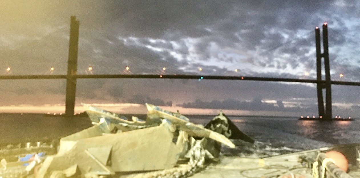 Large mud-streaked fluke anchor on a barge deck at dusk, with a cable-stayed bridge and a cloudy sky behind it