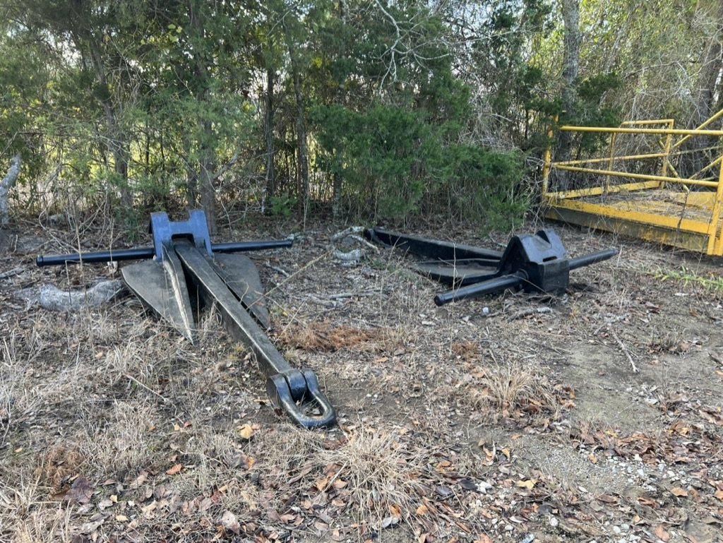 Two black stock anchors lying on dry grass and leaves in the yard beside a tree line