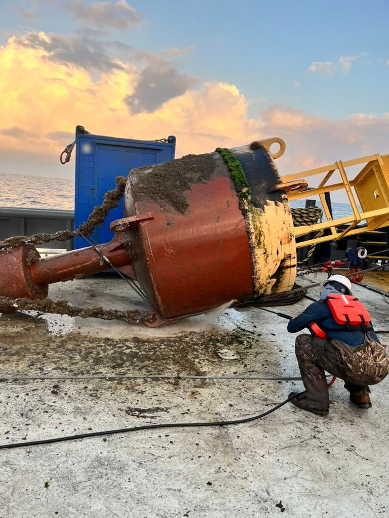 Crew member in a hard hat and life vest pressure-washing a large red and yellow buoy on a vessel deck at sea